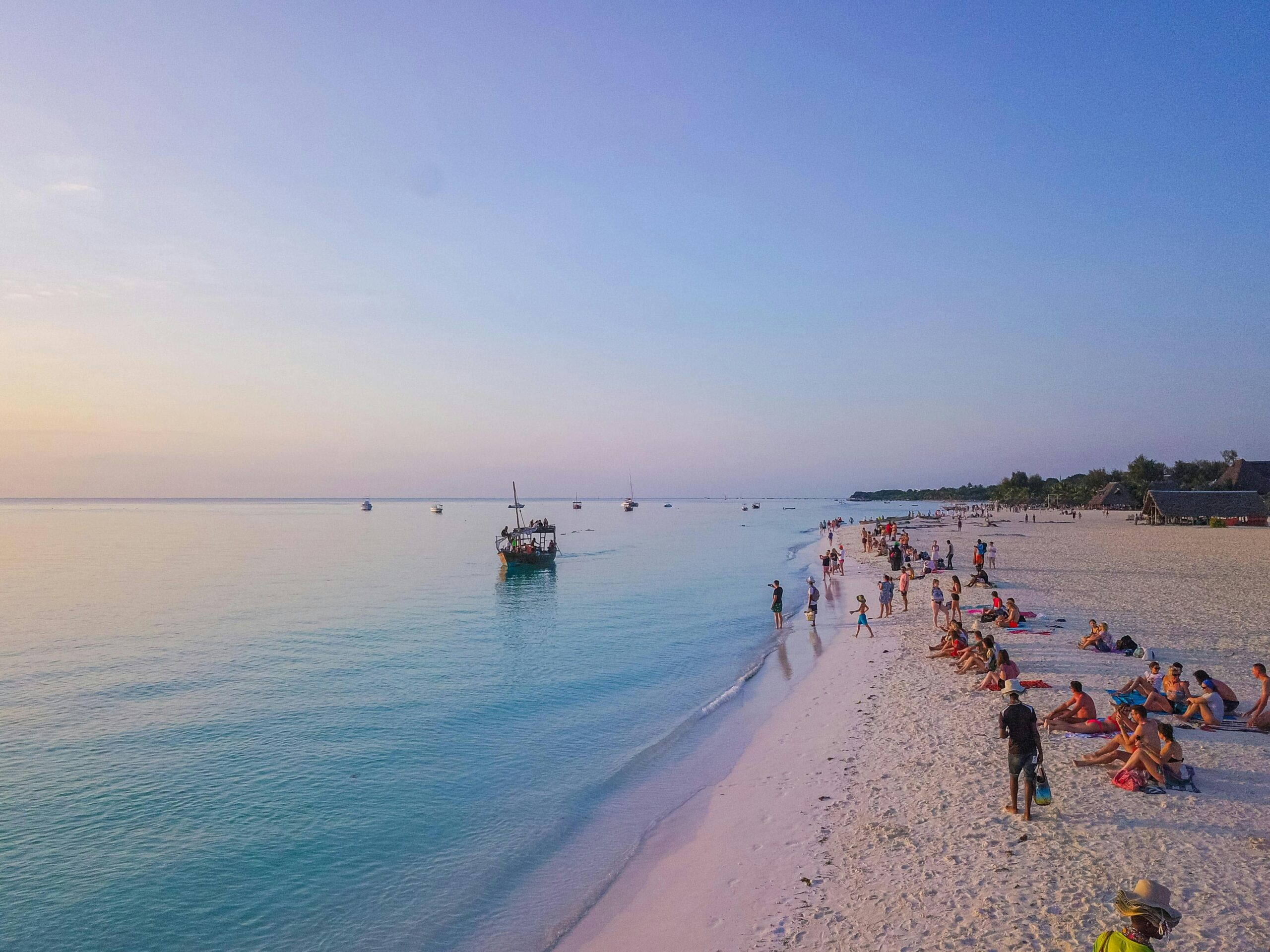 People enjoy a serene evening on a Tanzania beach with boats near the shore.