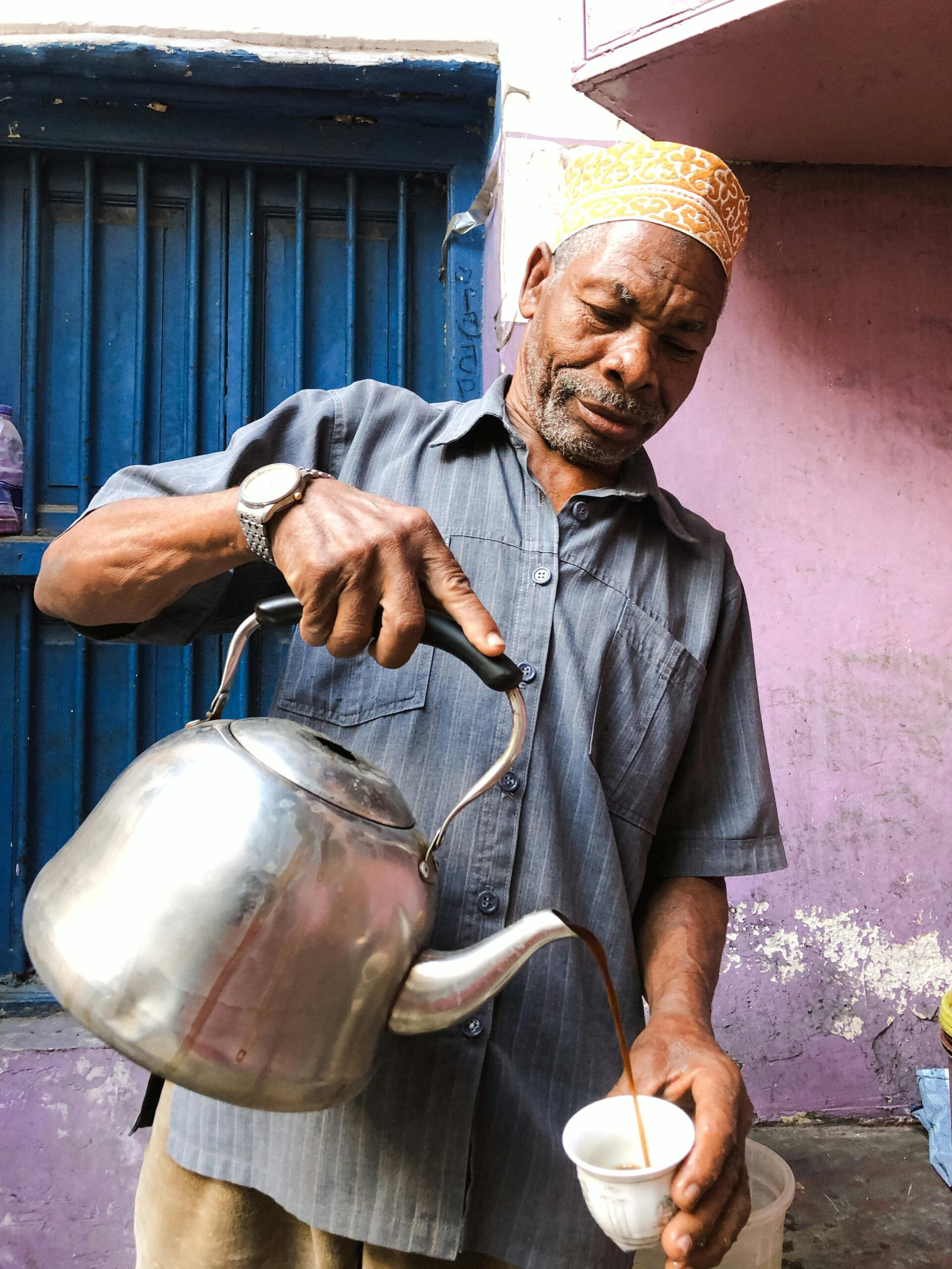 An elderly man pours coffee from a kettle on the streets of Tanzania. Vibrant colors frame the scene.