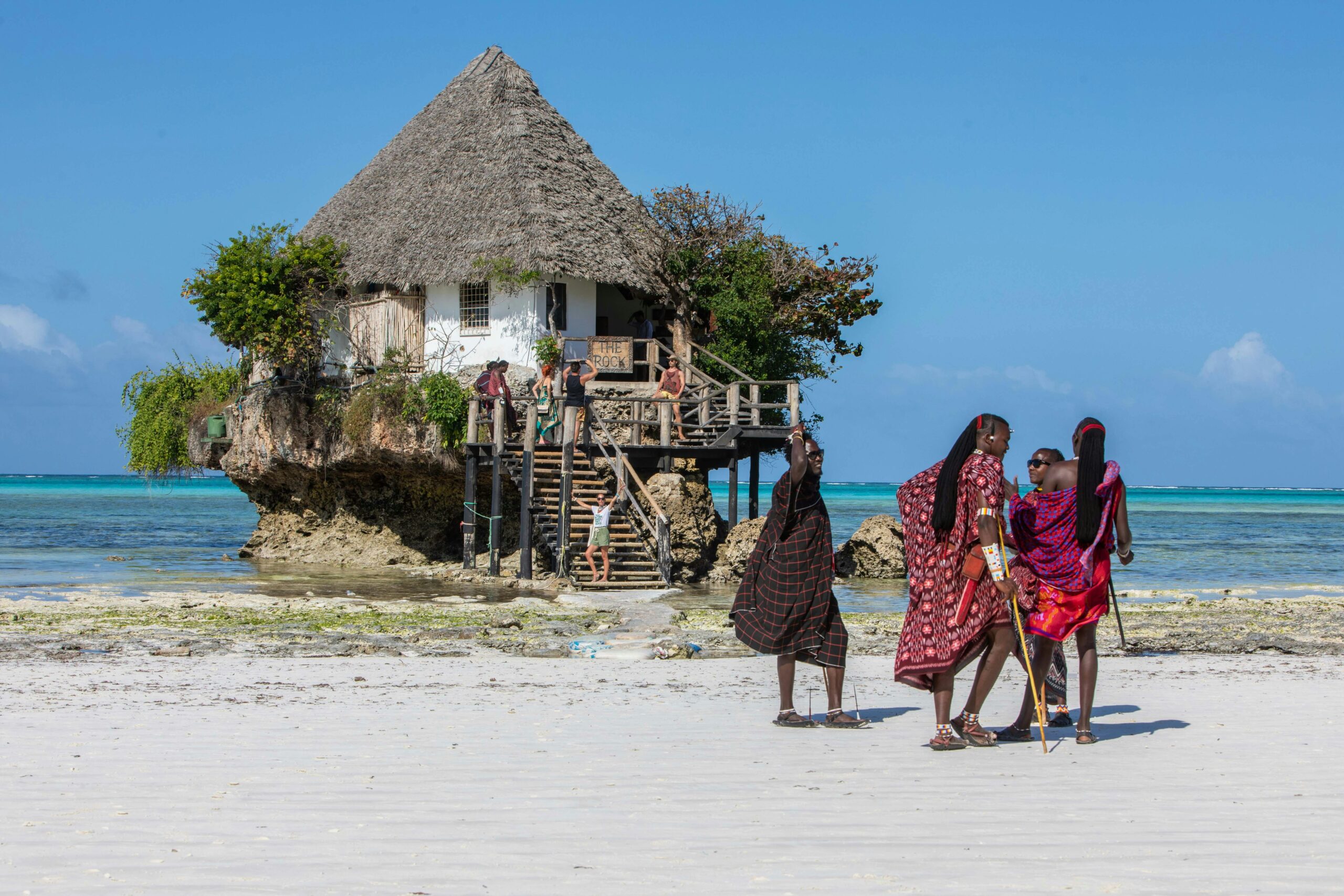 Cultural scene with The Rock restaurant and locals in traditional attire on a Zanzibar beach.