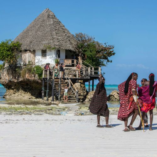 Cultural scene with The Rock restaurant and locals in traditional attire on a Zanzibar beach.